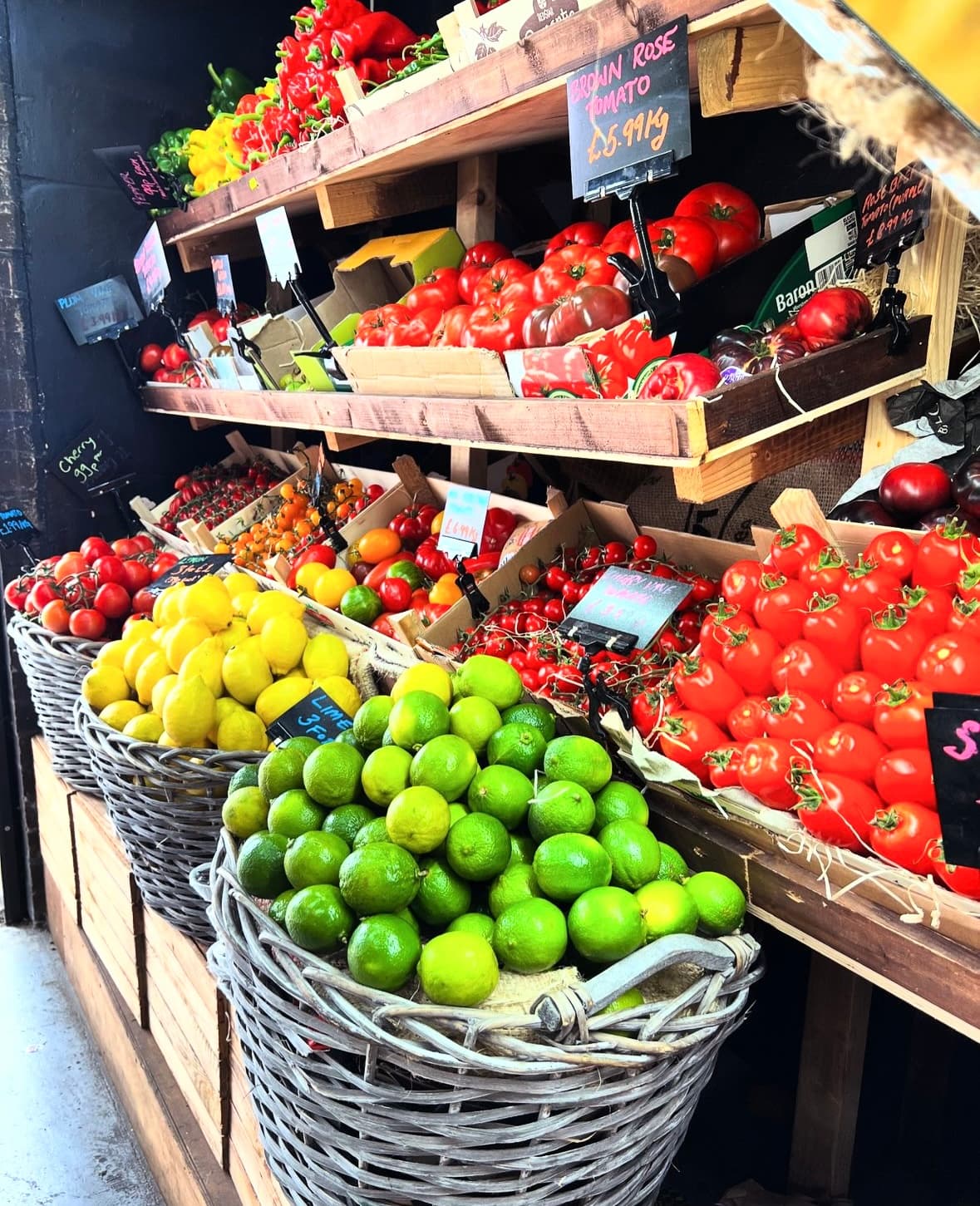 Wicker baskets of lemons and limes beside wooden crates of heirloom tomatoes at the Threadle produce wall, hand-written tags tied into the fruit.
